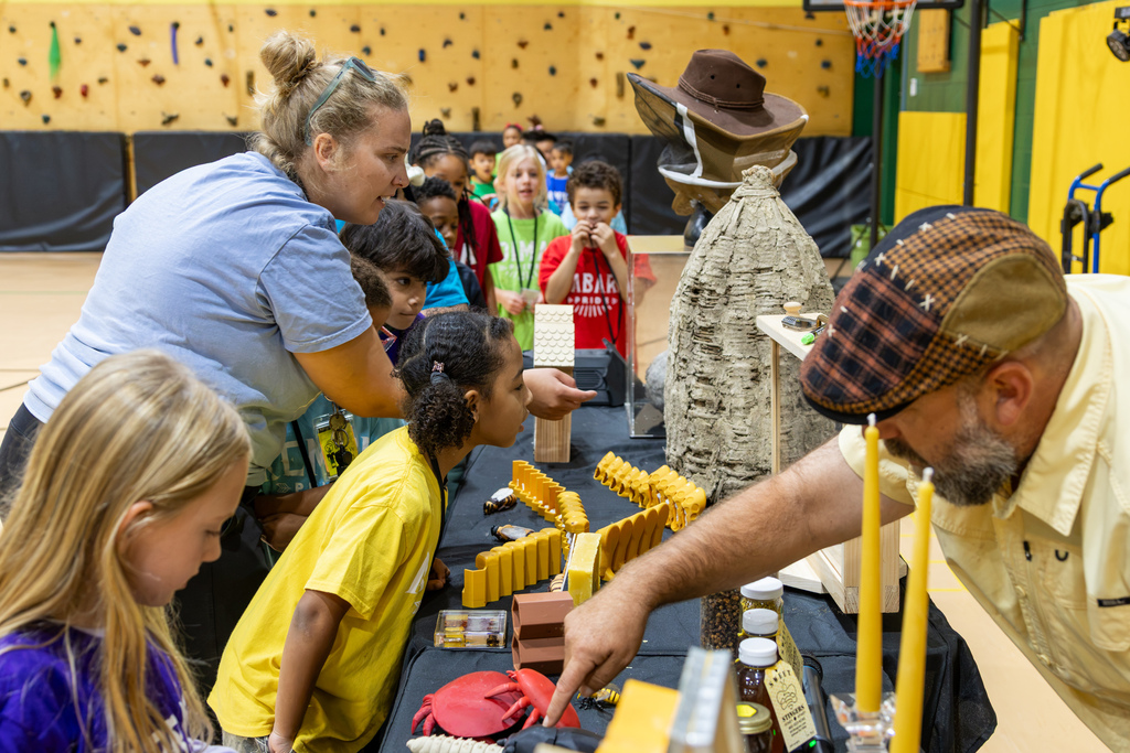 A presenter points out display items on a table while students and a teacher lean in to listen and observe.