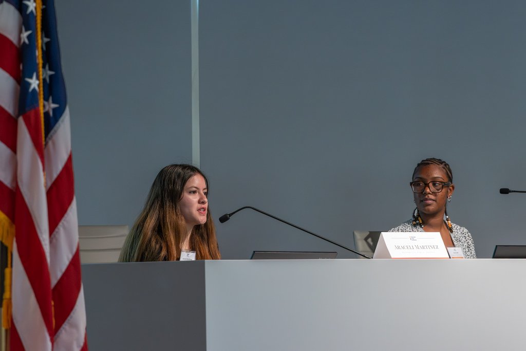 A female high school student panelist speaks into a microphone while sitting alongside a fellow student representative.