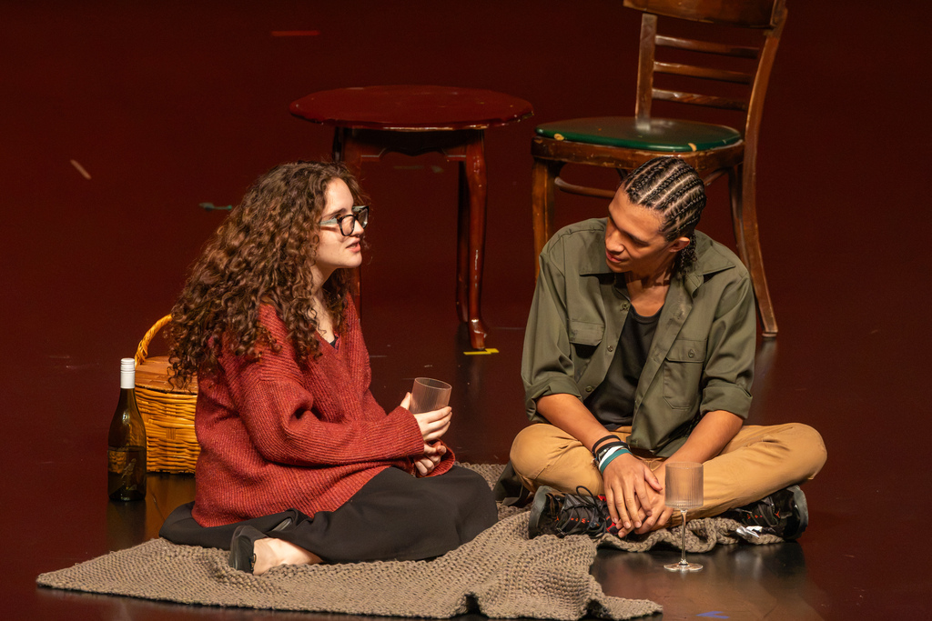 Two student actors sit together on a blanket on stage with a picnic basket prop during an intimate dialogue scene.