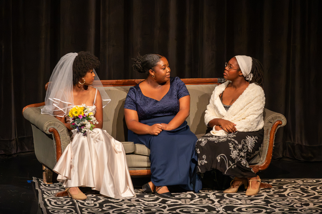 Three female student actors sit together on a vintage green couch on stage during a dramatic scene.