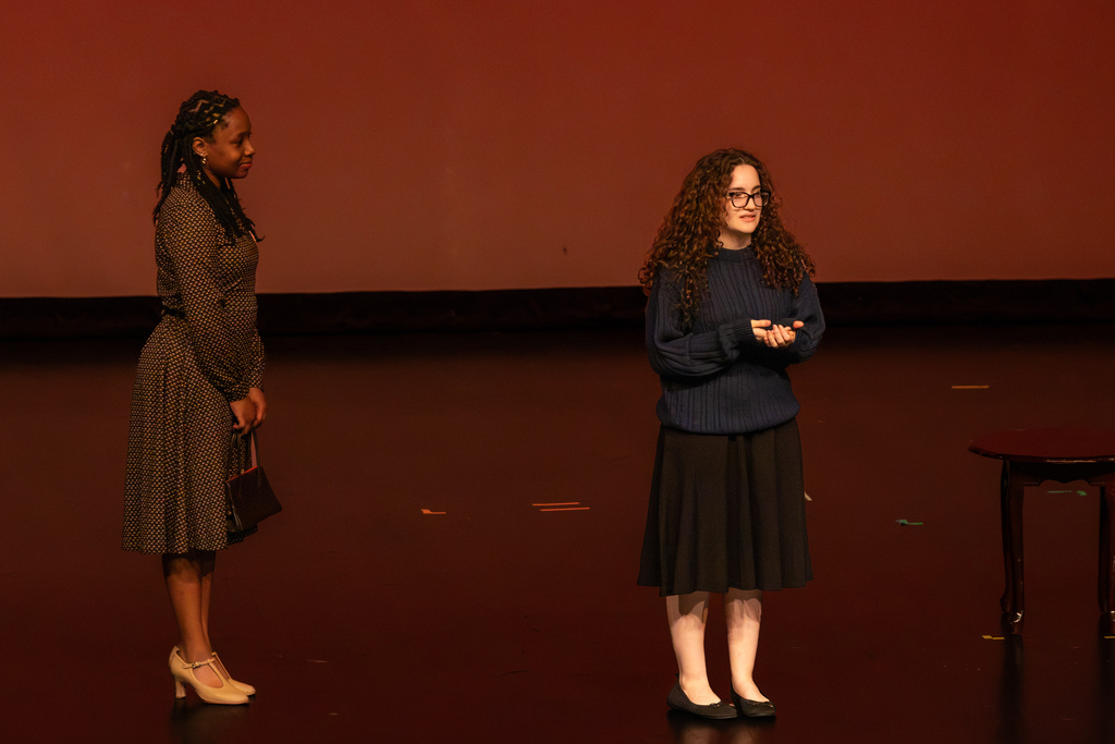 Two female student actors in retro-styled dresses speak and gesture during a performance on stage.