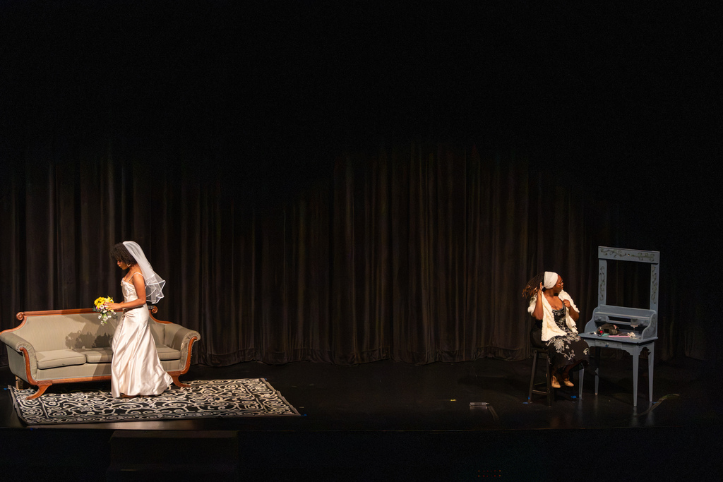 A student actor in a wedding dress and veil walks across the stage toward another performer seated at a vanity table.