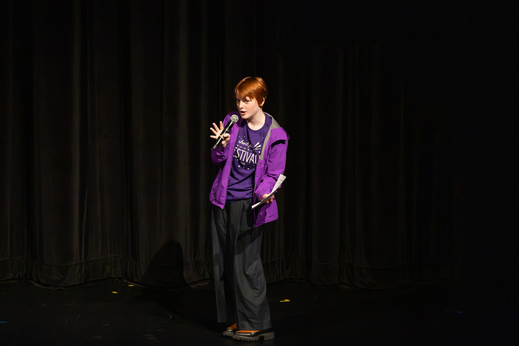 A student host wearing a purple "Short Play Festival" t-shirt and jacket speaks into a microphone on stage during introductory remarks.