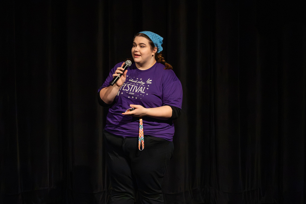 Mrs. Cook, PCH's theatre director, wearing a purple festival shirt and blue beanie speaks into a microphone to address the audience.