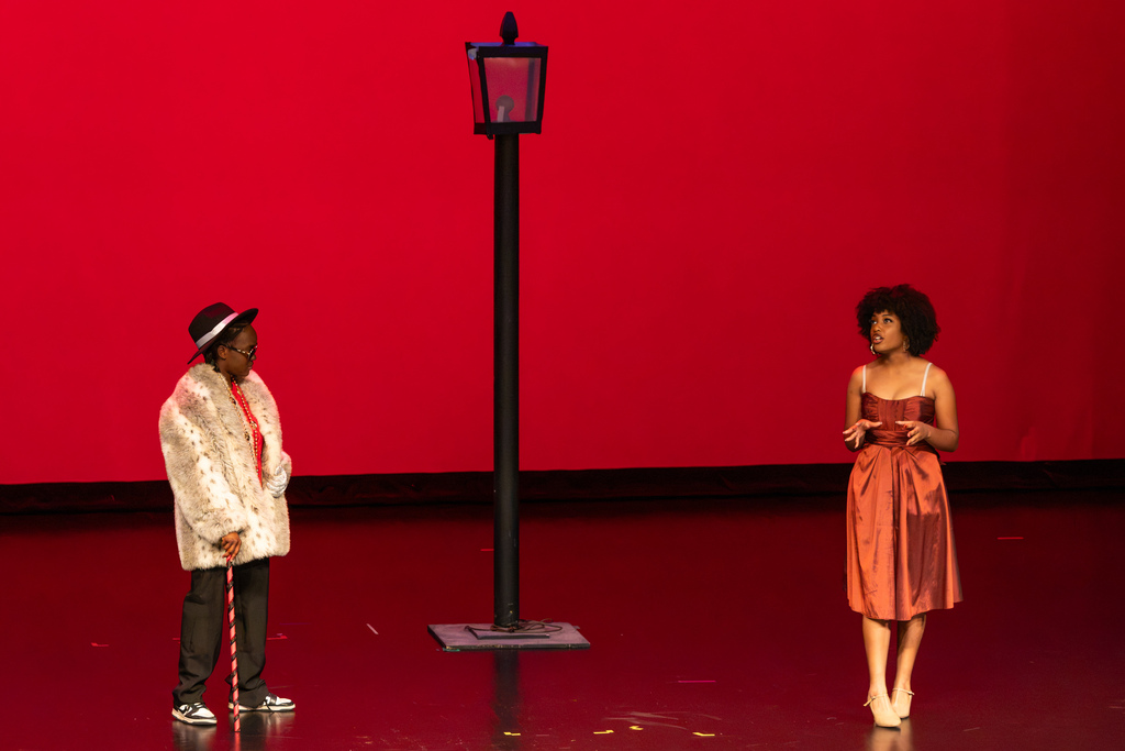 Two student actors perform a scene under a red stage light next to a streetlamp prop.