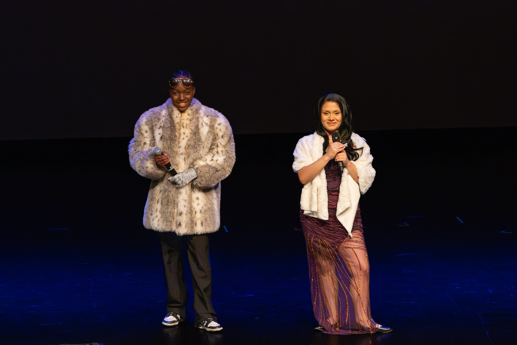Two student actors dressed in glamorous fur coats and formalwear hold microphones while performing on stage.