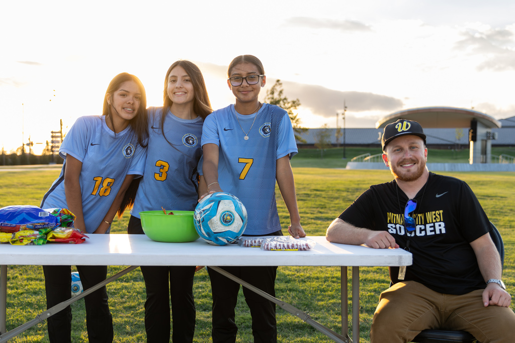 Three PC West girls soccer players and their coach smile behind a booth table stocked with candy and soccer balls.