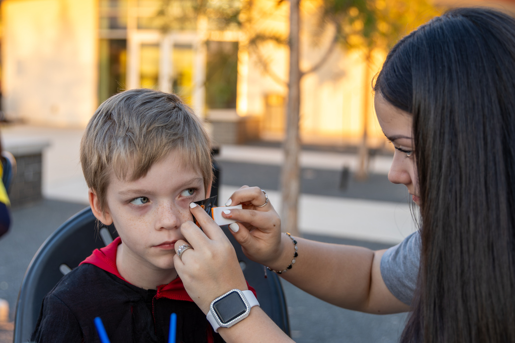 A high school volunteer applies a festive face stencil to a young boy's cheek.