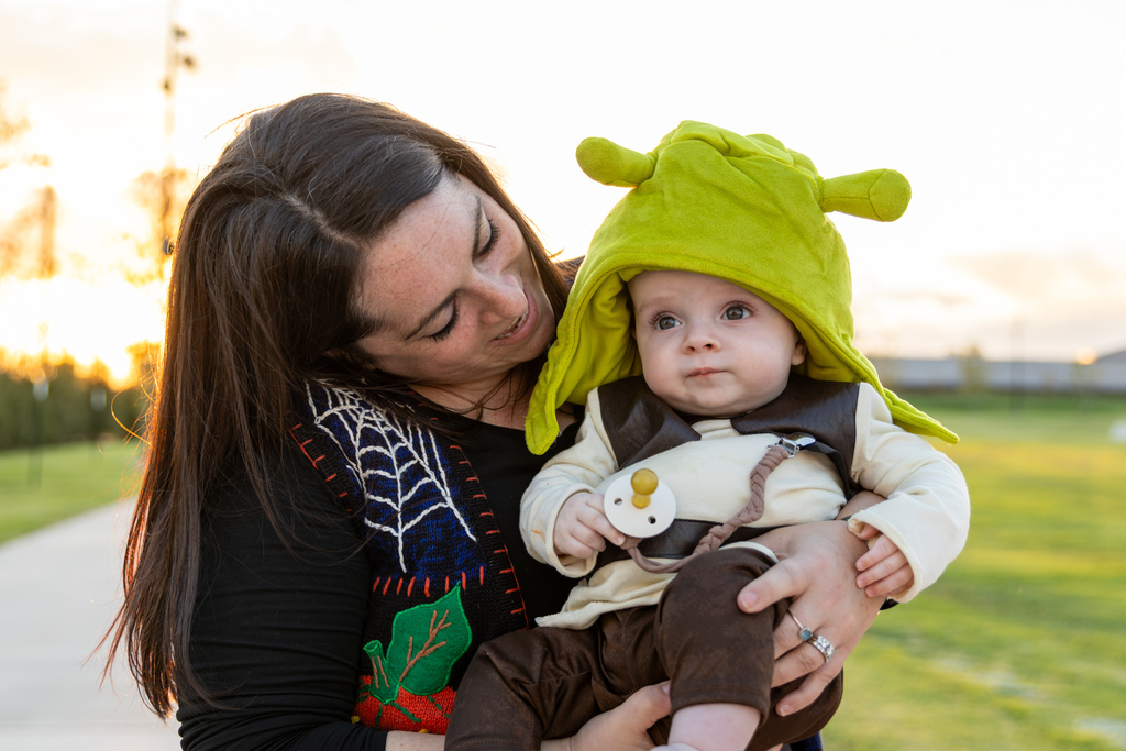A mother holds a baby dressed up in a green Shrek hoodie and costume.