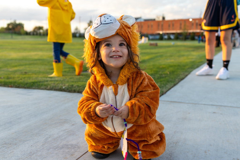 A toddler dressed in a furry lion costume crouches on the sidewalk and smiles up at the camera.