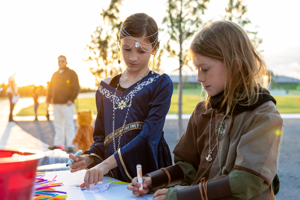 Two young students in medieval and Renaissance costumes lean over a craft table to color paper cutouts together.