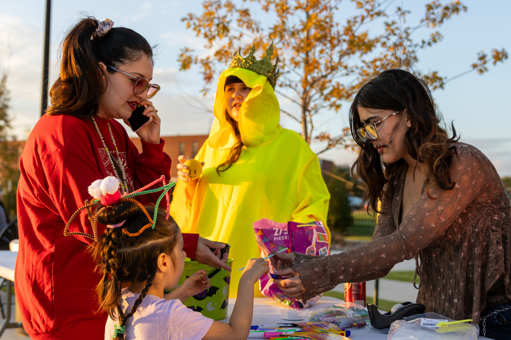 High school student volunteers hand out candy and crafts to trick-or-treaters at a festival table.