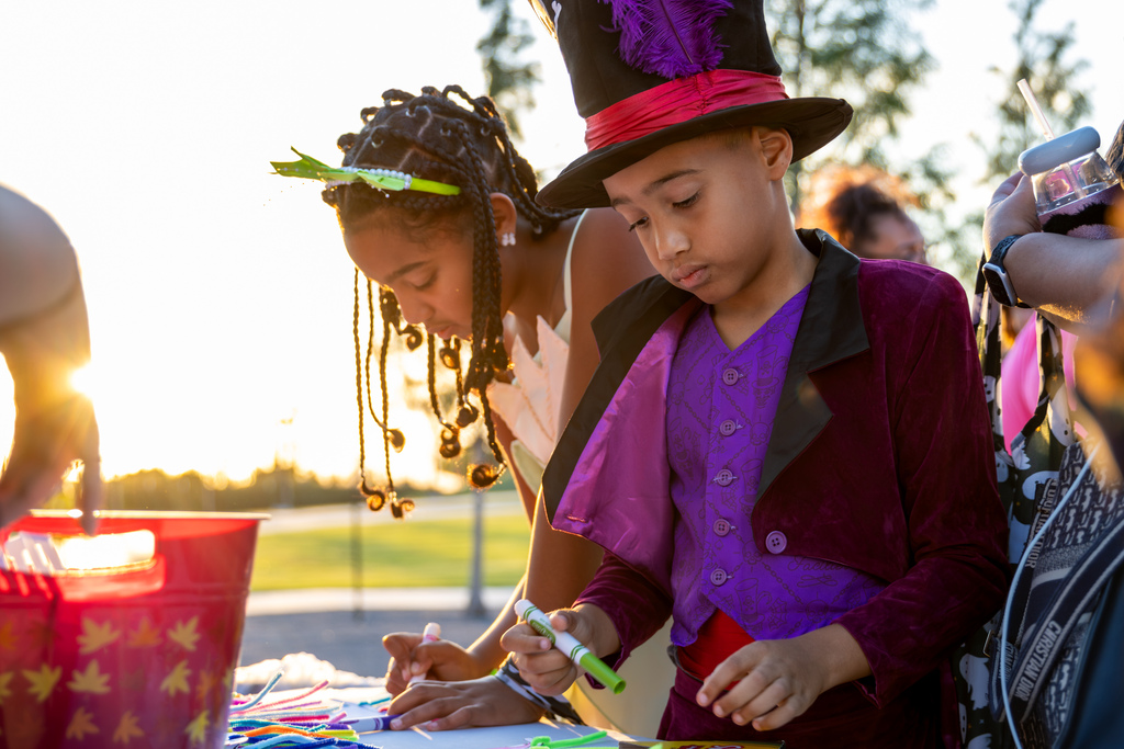 Two children in elaborate costumes—one in a top hat and purple vest—work intently on a craft project at an outdoor table at sunset.