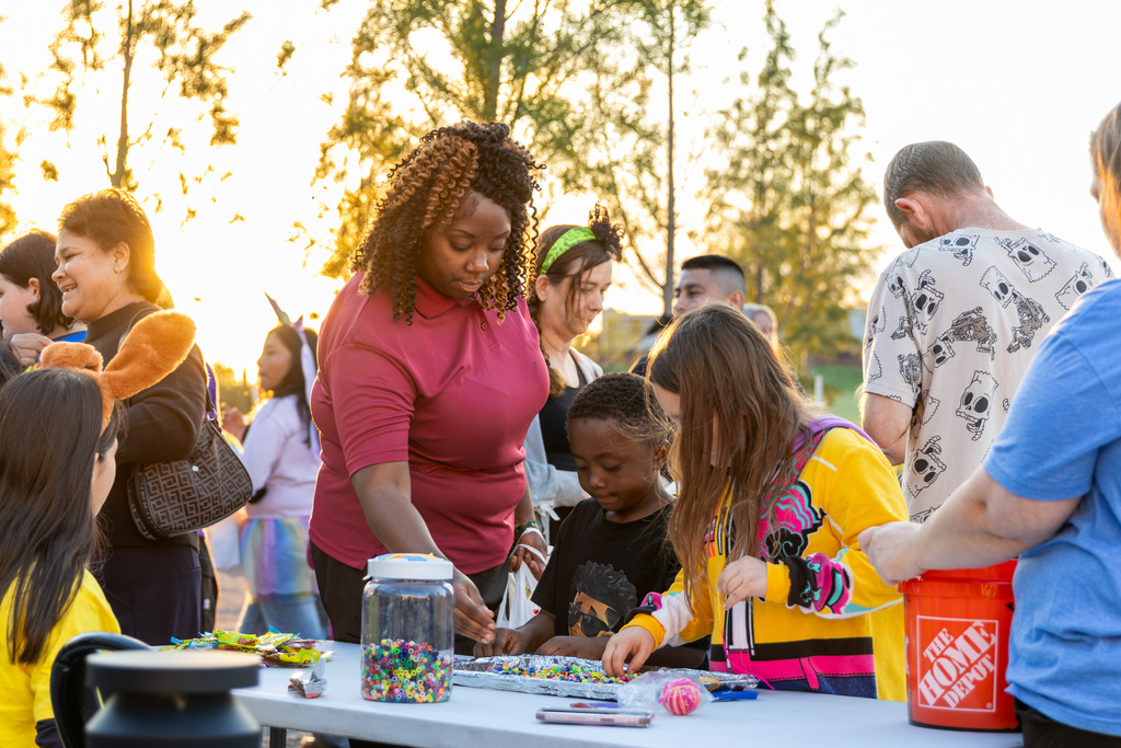 Volunteers assist young children with bead crafts and activities at a busy outdoor table.