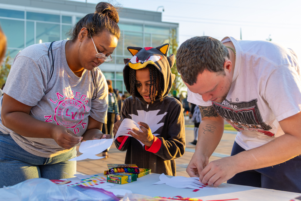 A child dressed in a Shadow the Hedgehog costume works on a paper craft at an outdoor table alongside two adults.