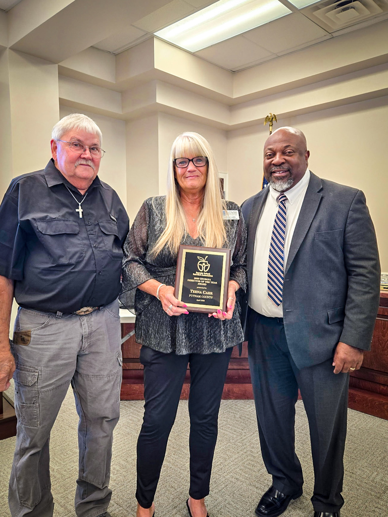 Three people in a room with a beige backdrop. One holds a plaque while the other two smile.