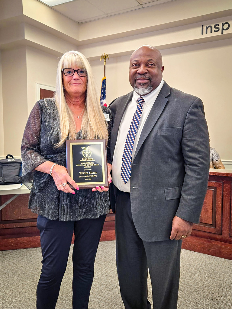 A woman with long blonde hair holds a plaque, standing next to a man in a suit. They stand before a podium.