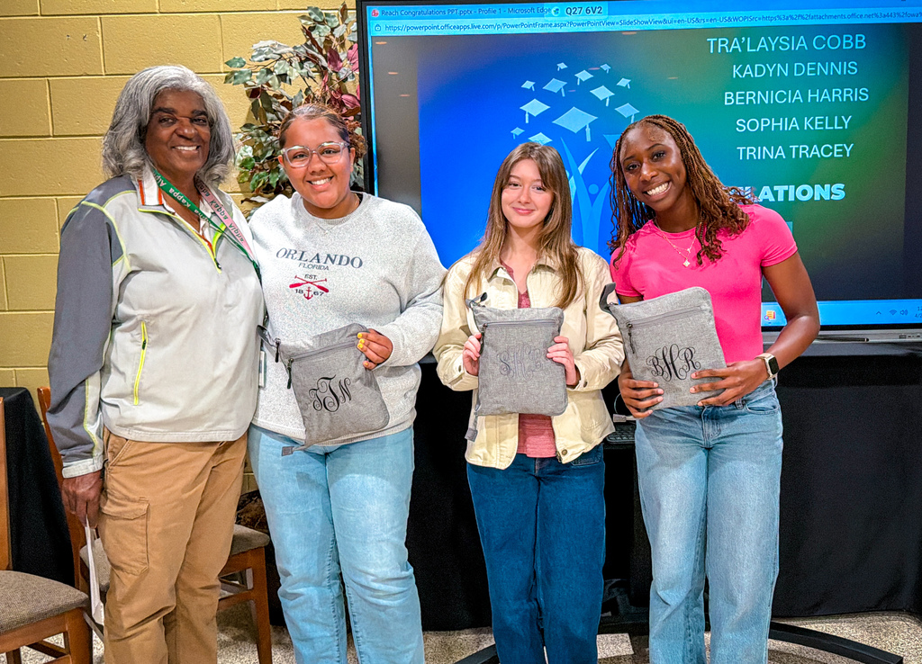 Four women standing with pillows in front of a large screen with a list of names.