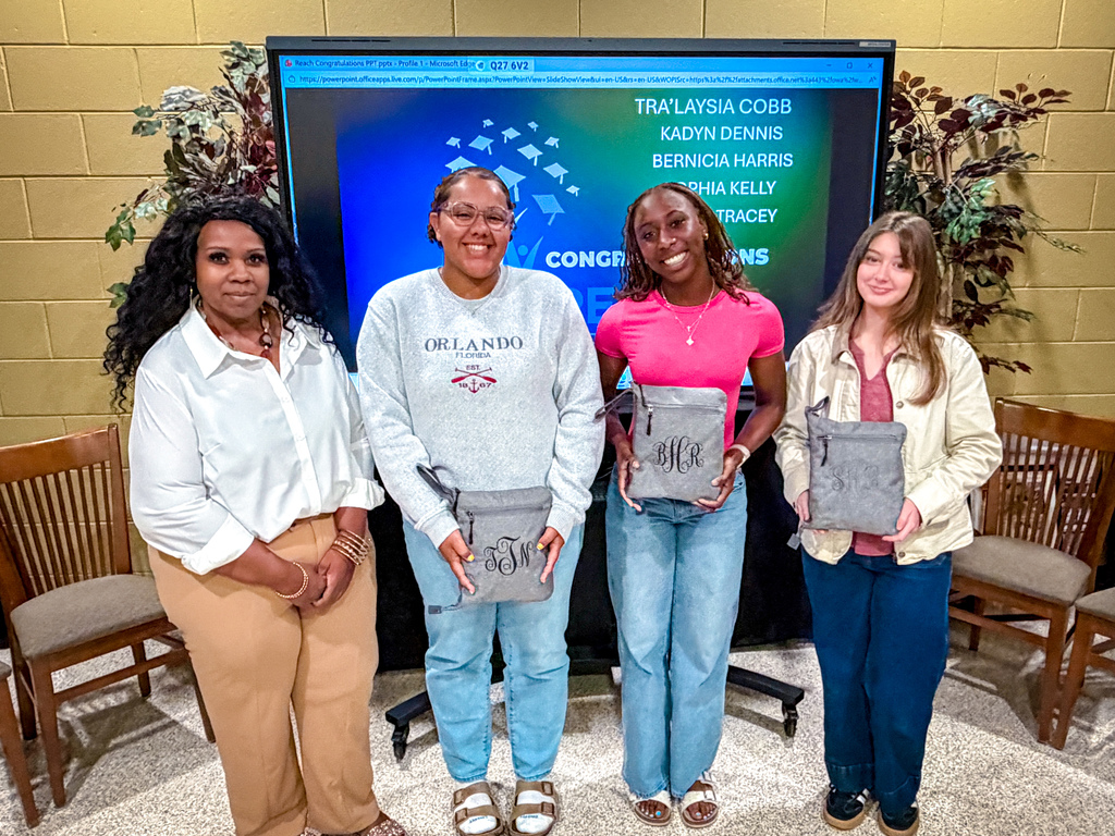 Four women stand in front of a monitor. They each hold a gray bag. Two of them wear white shirts.