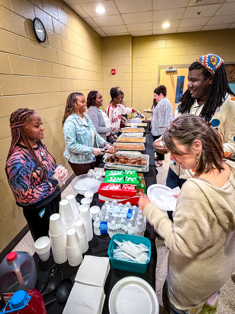 A group of people at a long table with food, cups, and bottles. The wall has a clock and yellow paint.
