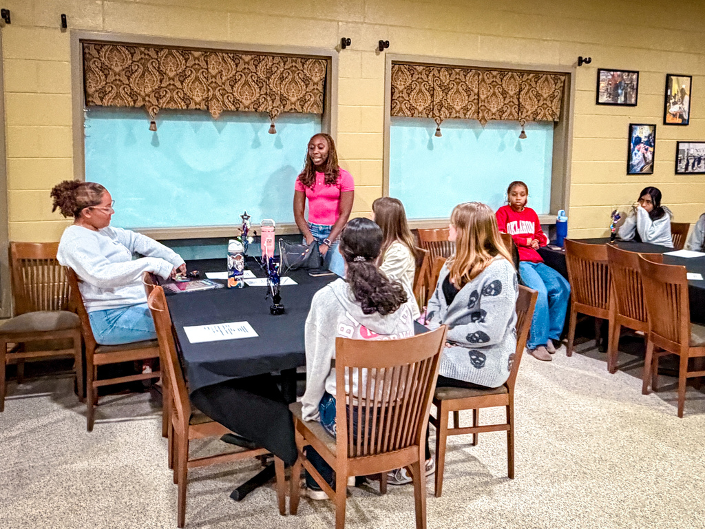 Group of people seated around a table in a room with beige walls, windows, and framed pictures.