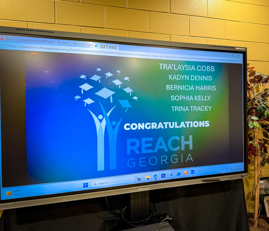 A monitor displays a blue screen with "Congratulations" text and names. A tree logo, "REACH Georgia" text, and icons are visible. The background features yellow walls and a potted plant.