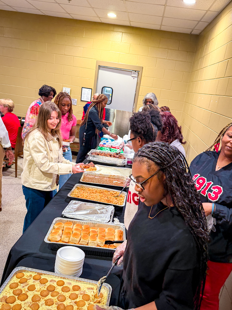 Several women are around a table, with trays of food. One person uses a spoon to serve food.