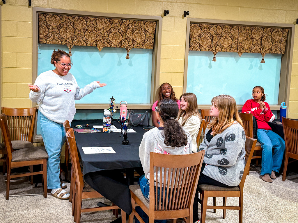A woman speaks to a group of girls seated around a table in a room with yellow walls and large windows.