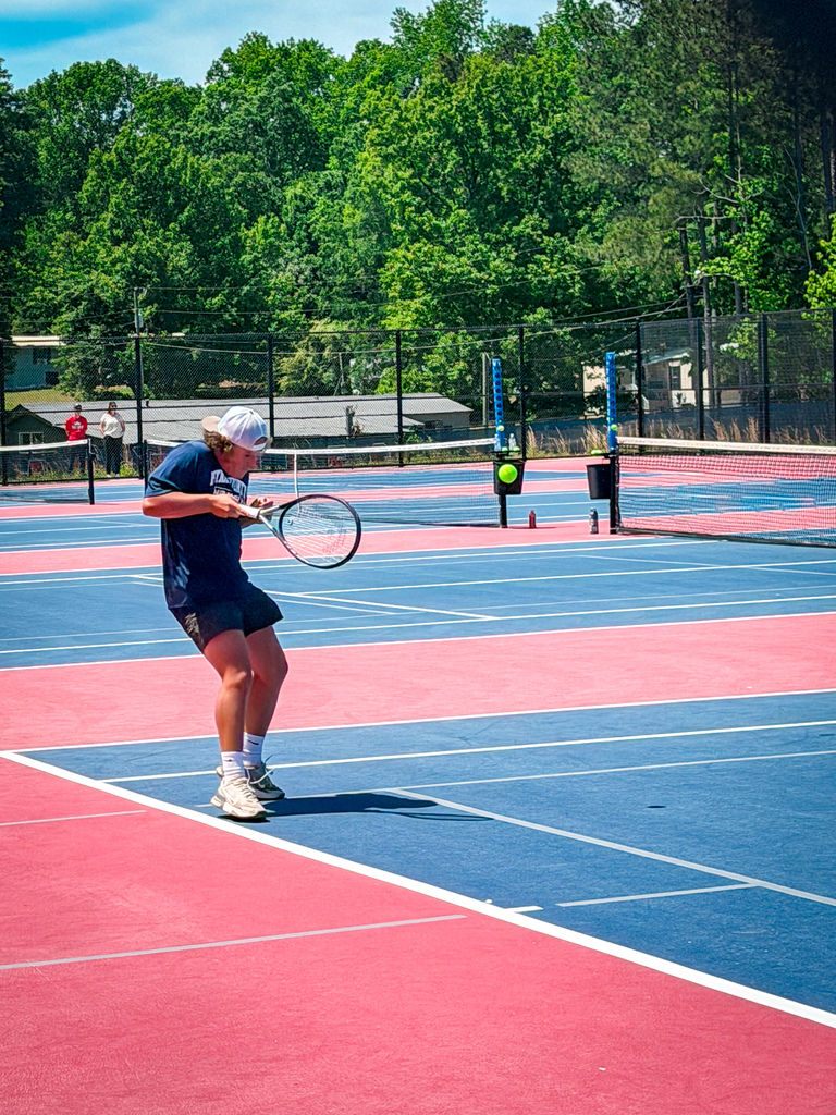 A man playing tennis on a blue and red court. Trees and a building in the background.