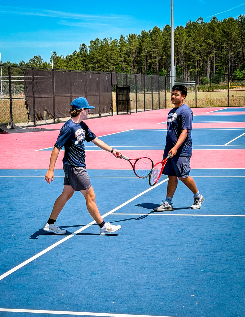 Two individuals playing tennis on a blue court. They are holding tennis rackets. Behind them is a fence.