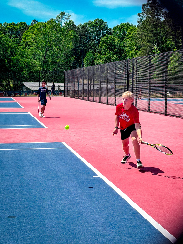 Two people playing tennis on an outdoor court, one wearing a red shirt and the other a black shirt.