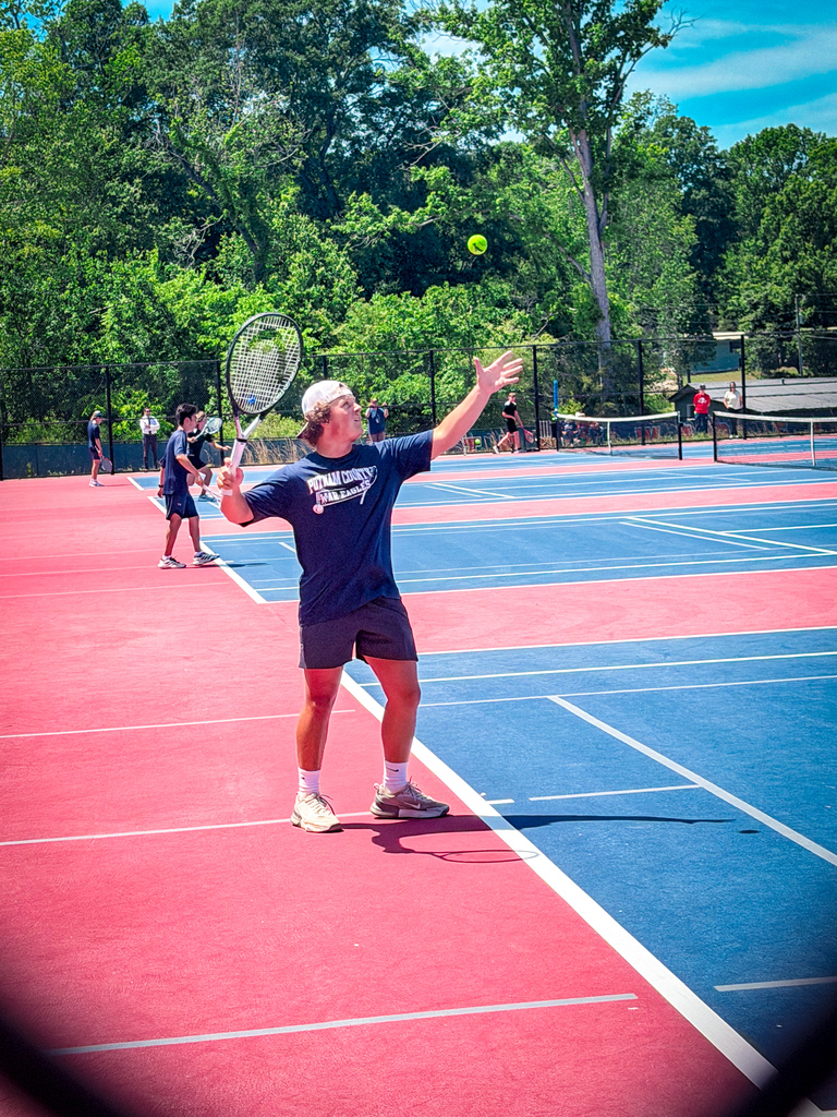 A tennis player in blue shirt and shorts swings a racket with a yellow ball above. Red and blue tennis court with other players.