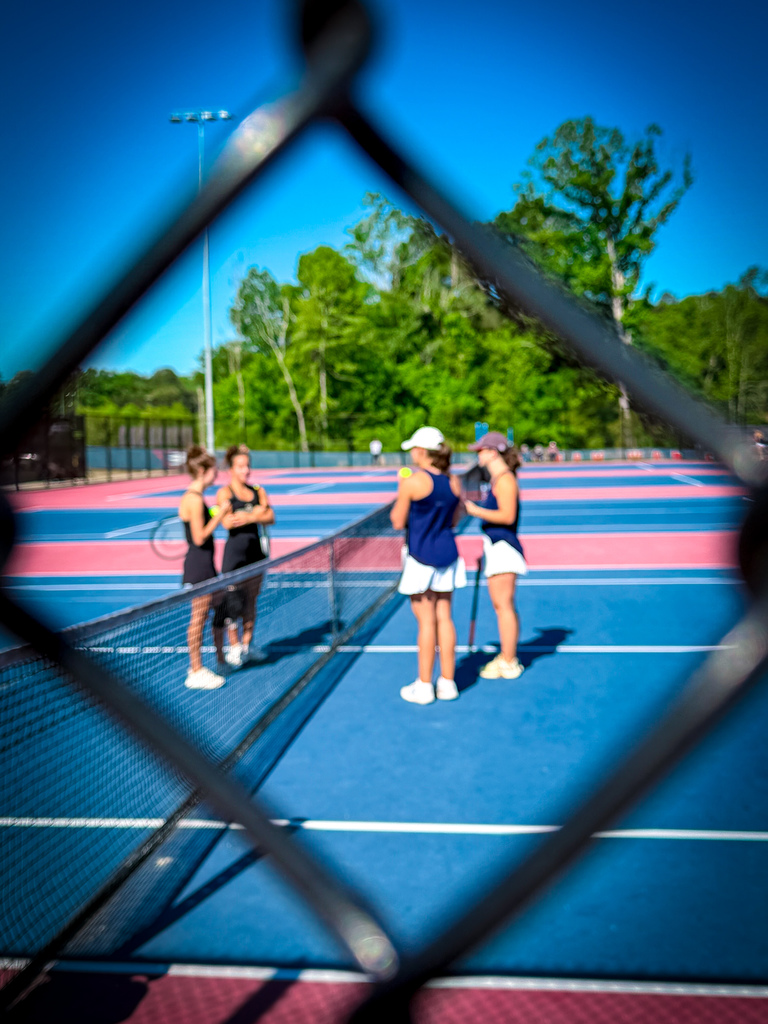 Four women stand on a tennis court with blue and pink markings. Two hold rackets, one wears a hat, and the court is surrounded by trees.