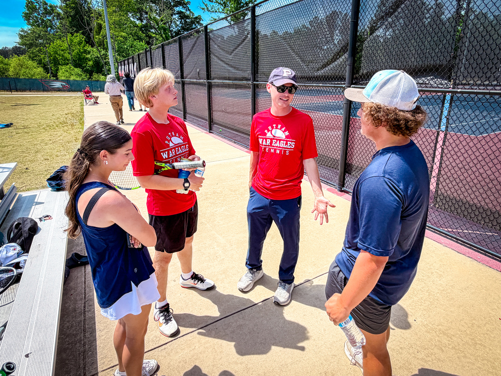 Four individuals converse on a tennis court with a black fence. Two wear red shirts, and two wear blue shirts.
