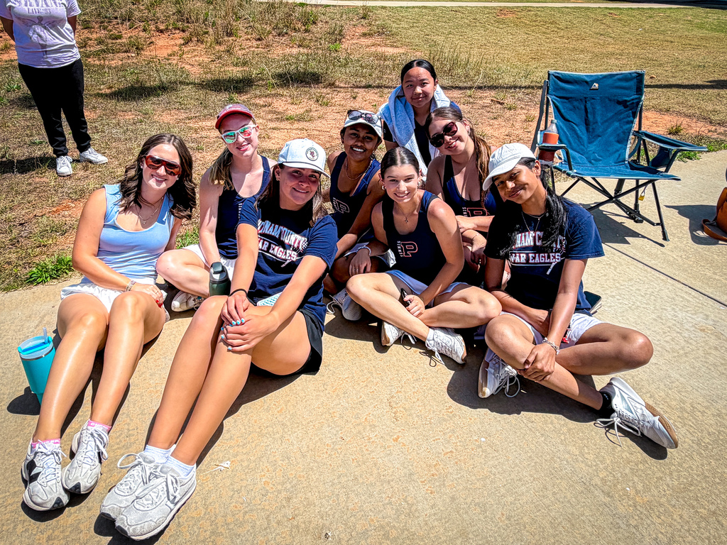 Group of women in athletic wear sitting on a concrete path with dry grass and a blue chair in the background.