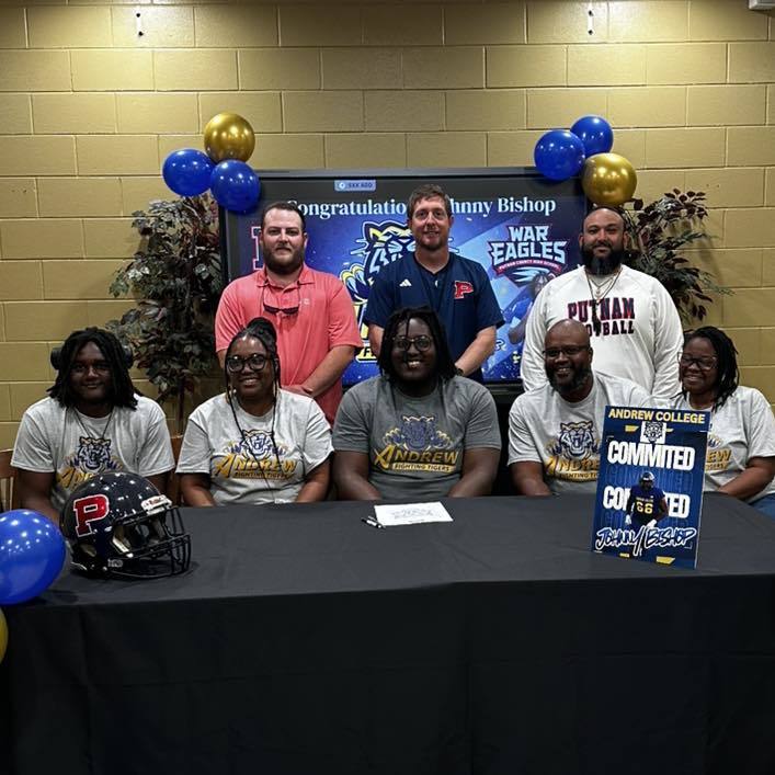 Nine individuals pose at a desk, smiling. One holds a football helmet. Behind them, balloons and a sign read "Congratulations."