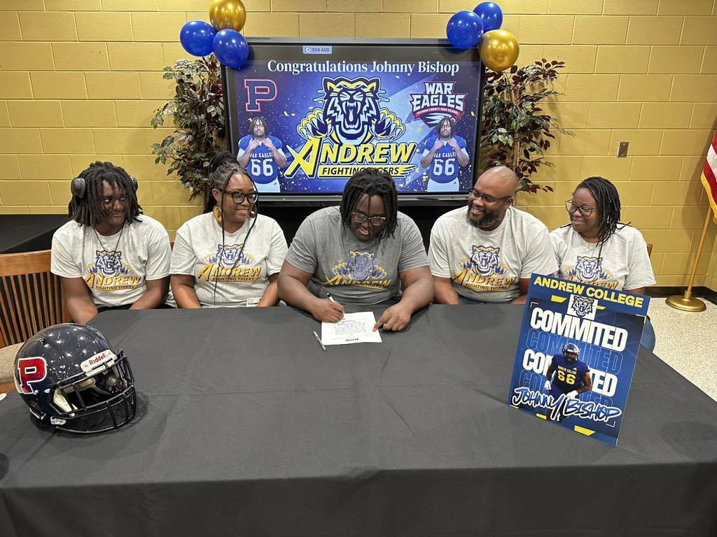 Five men sit at a table, signing papers. One wears a football helmet. Behind them, a large screen says "ANDREW" and "56".
