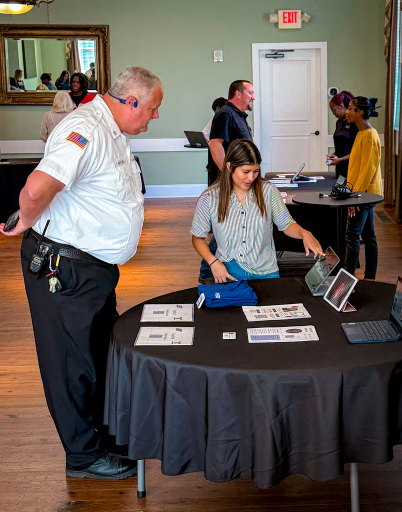 People in a room with a table displaying brochures, laptops, and a bag. A man in uniform stands near the table.