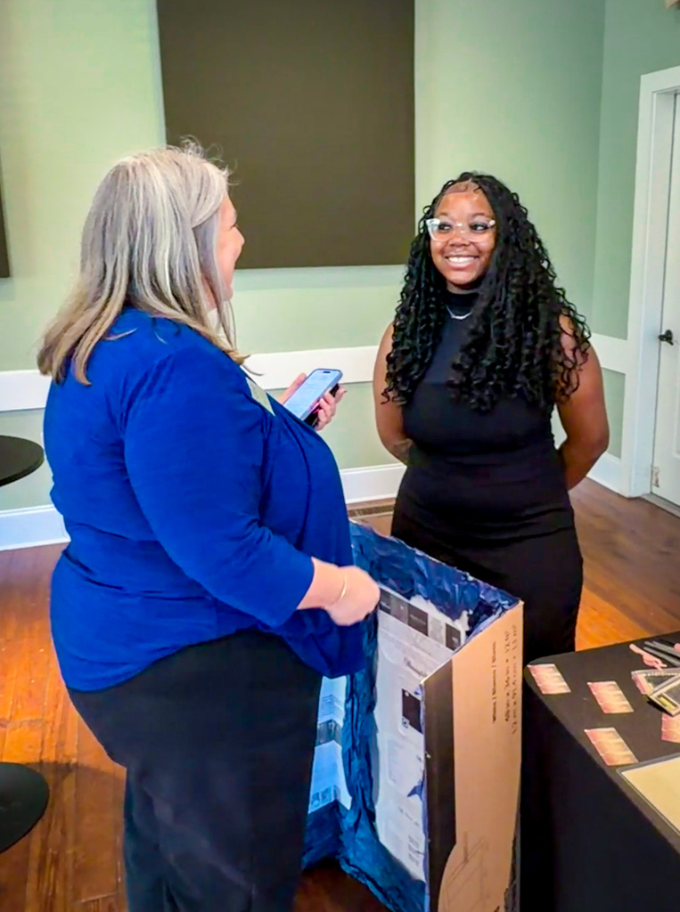 Two women, one holding a box and speaking to the other, standing in a room with a table and green walls.
