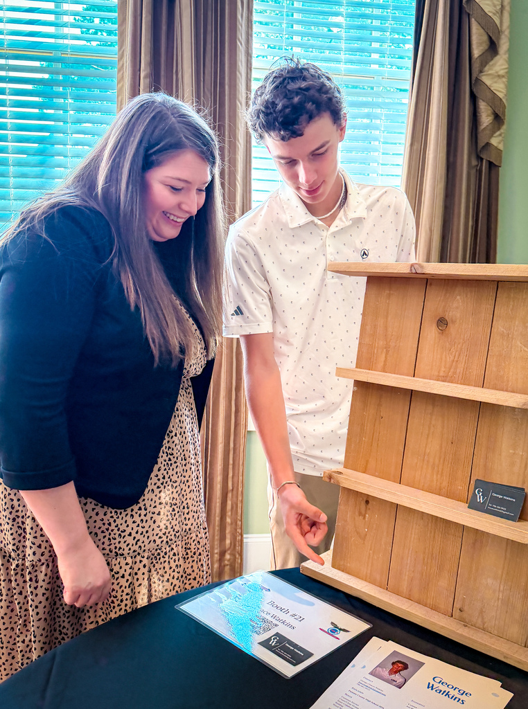 A woman and a boy examine a wooden shelf and brochures on a table in a room with windows.