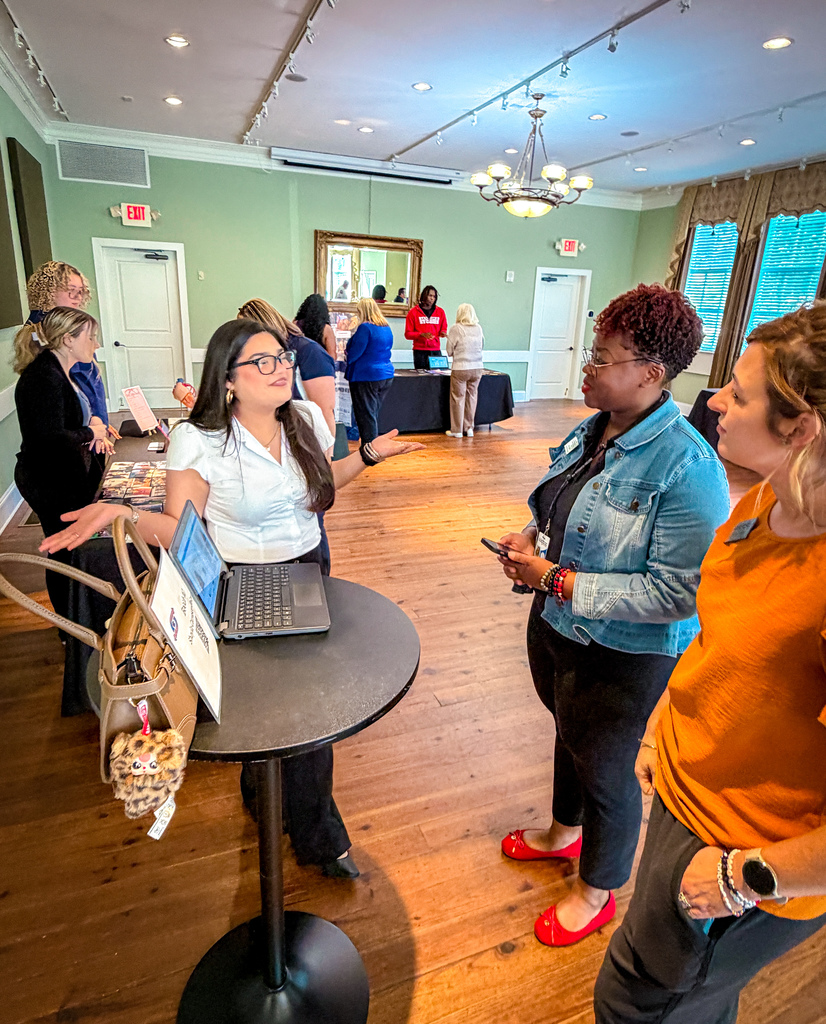 Several women in a room with wooden floors, one looks at a laptop on a table.