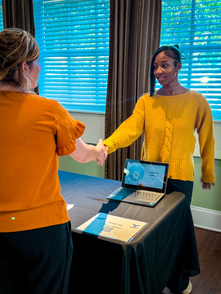 Two women are shaking hands in front of a table. One woman wears a yellow sweater, the other an orange top. A laptop with a screen is on the table.