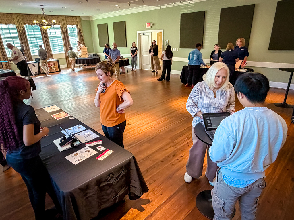 People in a room with hardwood floors gather around tables. Some wear casual clothing. One person uses a tablet.