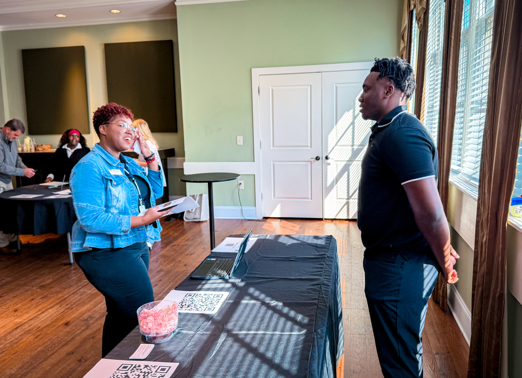 Two people stand by a table with a laptop and papers. One holds a piece of paper, the other stands with hands behind back.