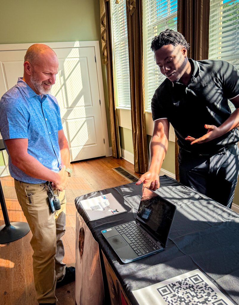Two men standing, one gesturing to a laptop on a table. One man in blue shirt and khakis, the other in black shirt and black pants.