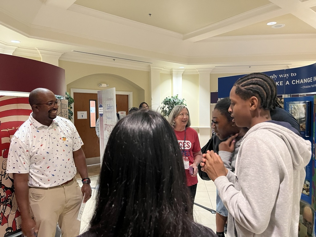 People standing in a hallway, engaging in conversation. One man wears glasses, a white shirt, and khaki pants.