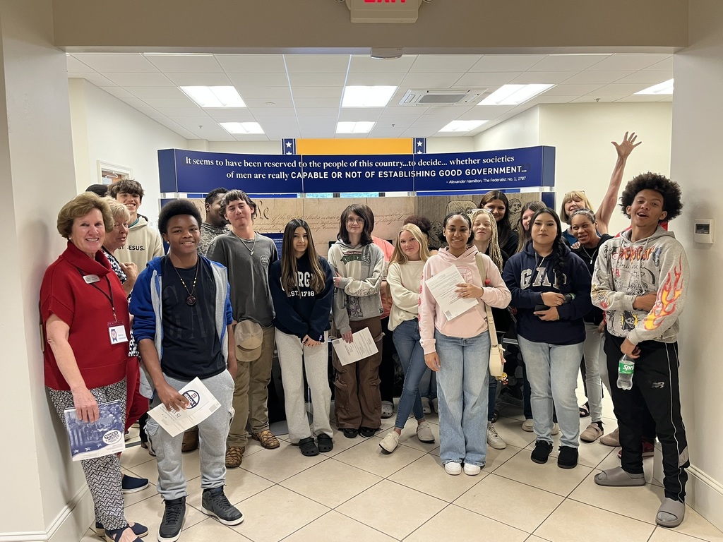 A group of young people stands together in a hallway, holding papers and smiling. A blue banner with text is visible above them.