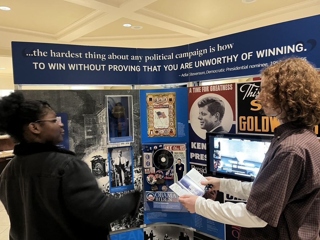 Two people in a room, one holding a paper, stand before an exhibit featuring photos and text.