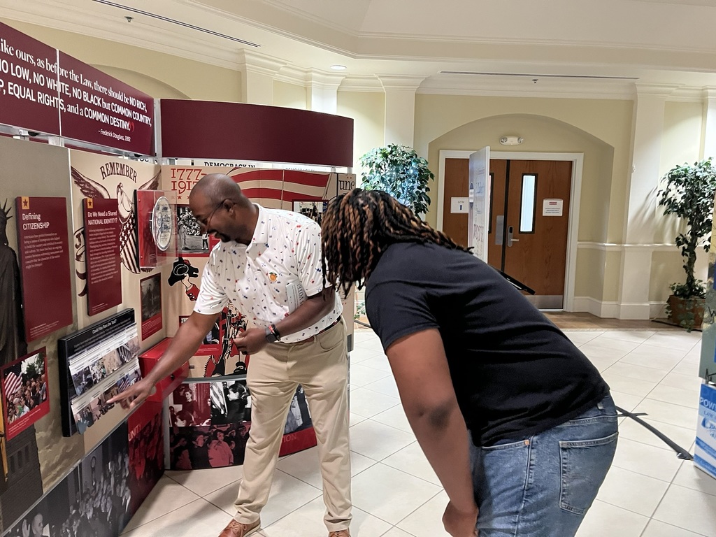 Two people examine a display in a museum exhibit. A man points at a photo. The exhibit has a red background.