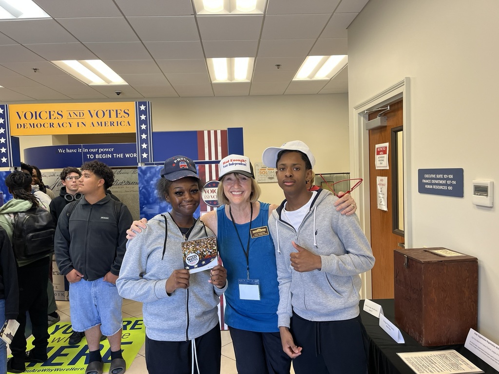 Three people stand together, smiling, near a table. One holds a trophy. Behind them, banners with "Voices and Votes" text are displayed.